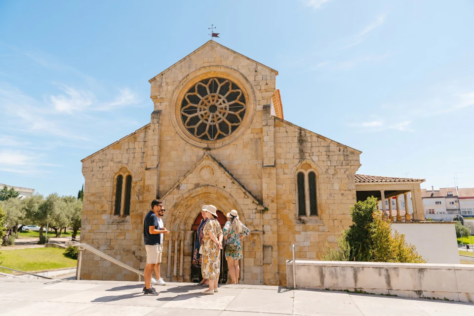Four people stand outside a stone church with a circular stained glass window and arched entrance on a sunny day.