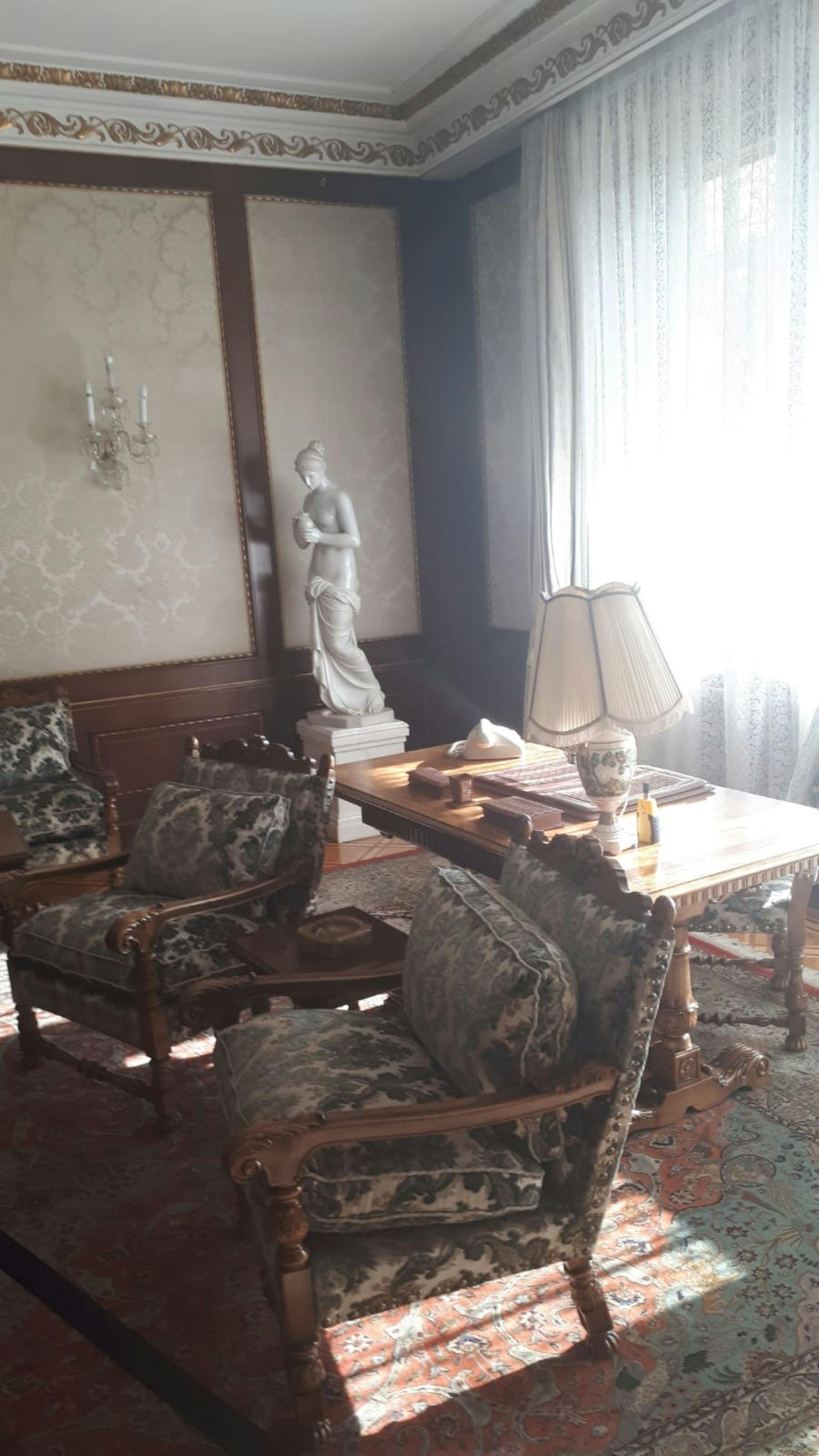 Una habitació amb mobles antics, un escriptori gran, cadires adornades, una estàtua, un llum i llum que es filtra a través de cortines de encaix.