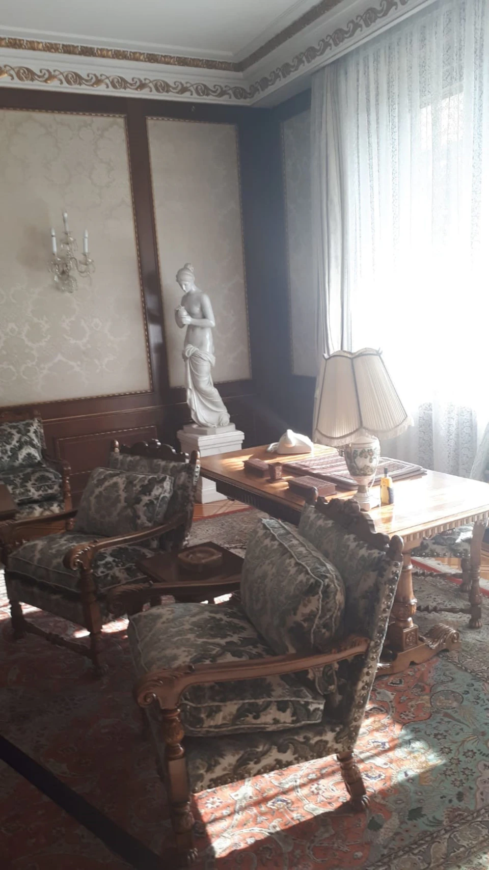 A room with antique furniture, a large desk, ornate chairs, a statue, a lamp, and light filtering through lace curtains.