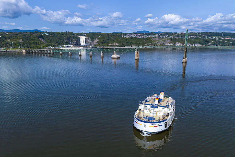 A passenger boat on a calm river with a waterfall, bridge, and hills in the background under a partly cloudy sky.