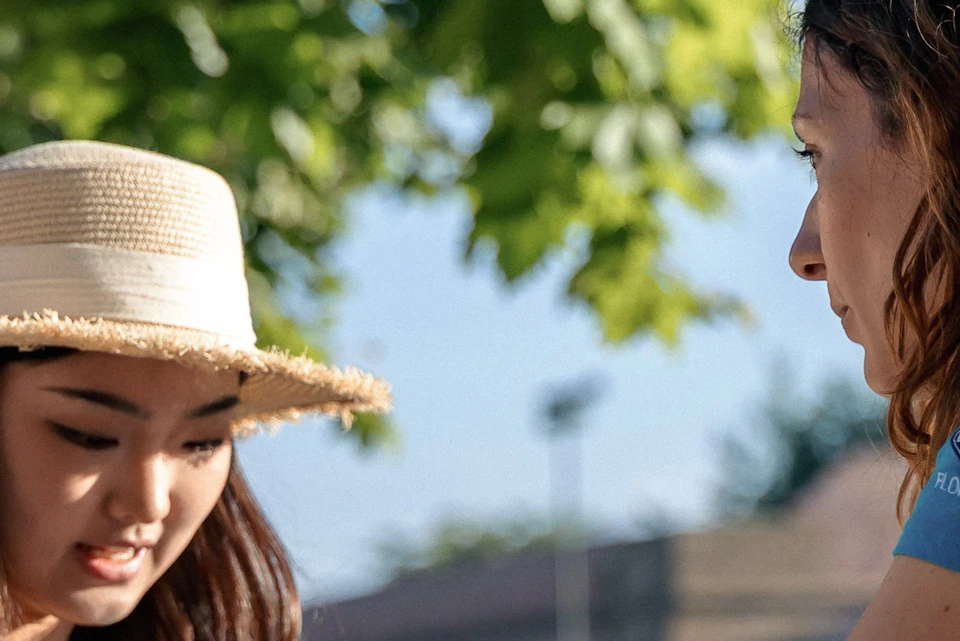 Two women interacting outdoors; one is handing an item to the other. A camera hangs around the neck of one woman.
