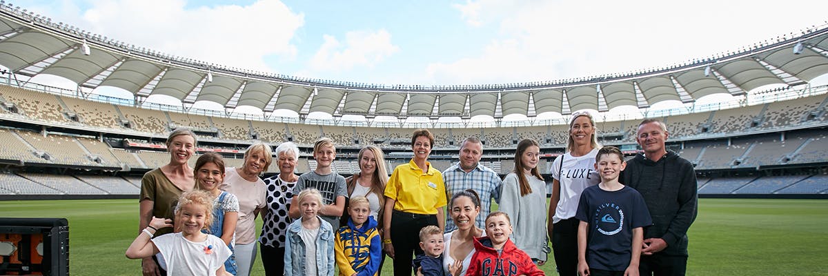 Μια ομάδα ενηλίκων και παιδιών στέκονται και χαμογελούν στο γήπεδο ενός μεγάλου, ως επί το πλείστον άδειου σταδίου κάτω από έναν γαλάζιο ουρανό με σύννεφα.