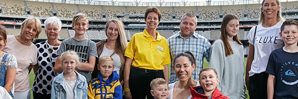 A group of adults and children stand and smile on the field of a large, mostly empty stadium under a blue sky with clouds.