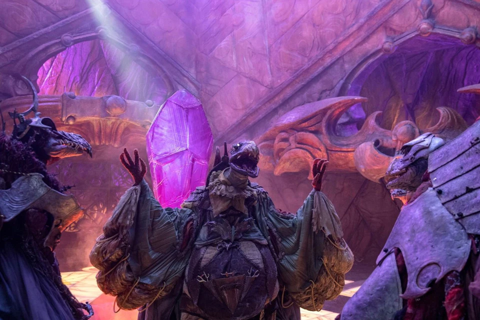 A detailed puppet creature in ornate robes, lit by purple light, standing in an elaborate fantasy set with carved decorations.