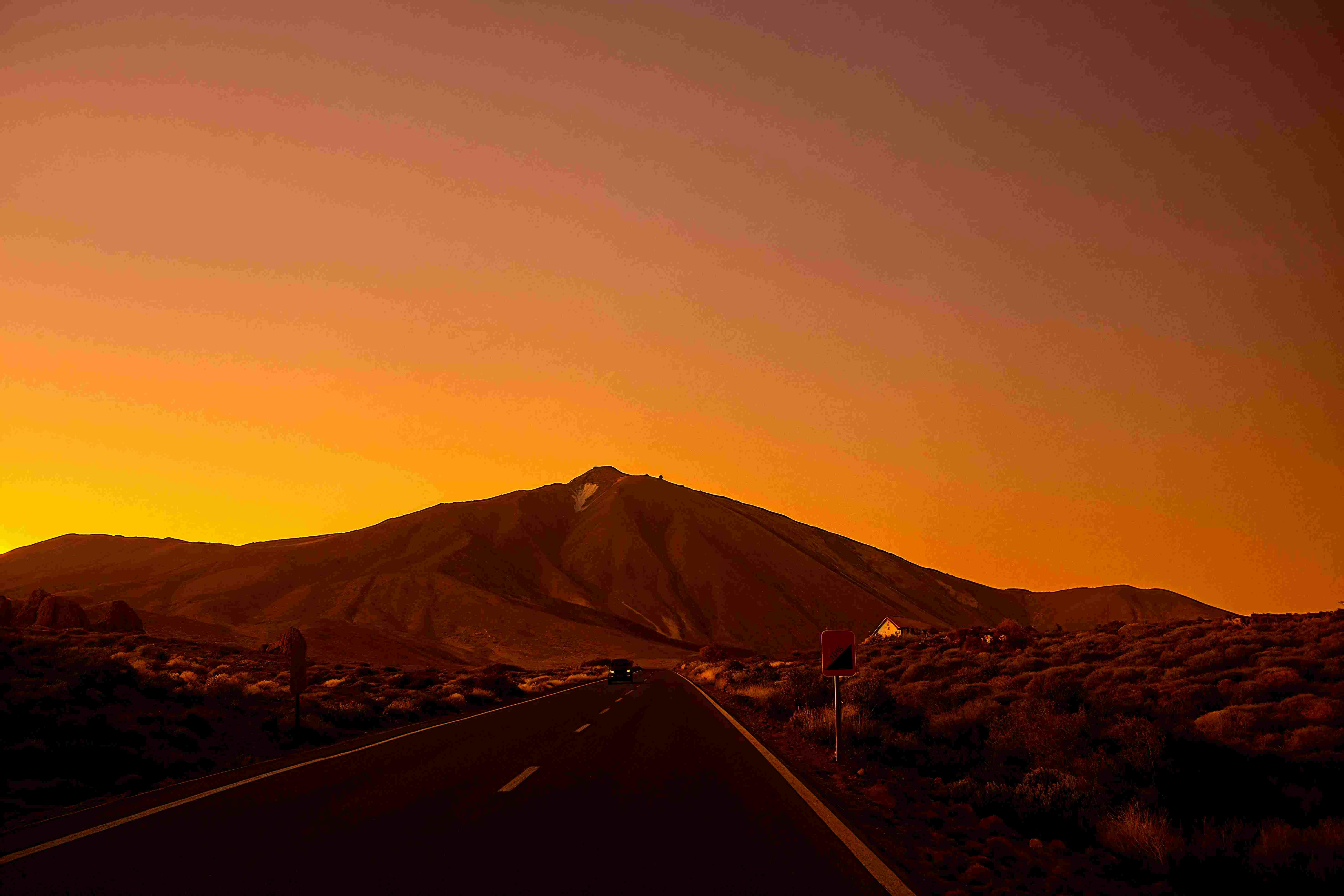 鮮やかなオレンジ色の夕焼けの空の下、道路に沿って山に向かって車が走っています。道路沿いには標識とまばらな植物があります。