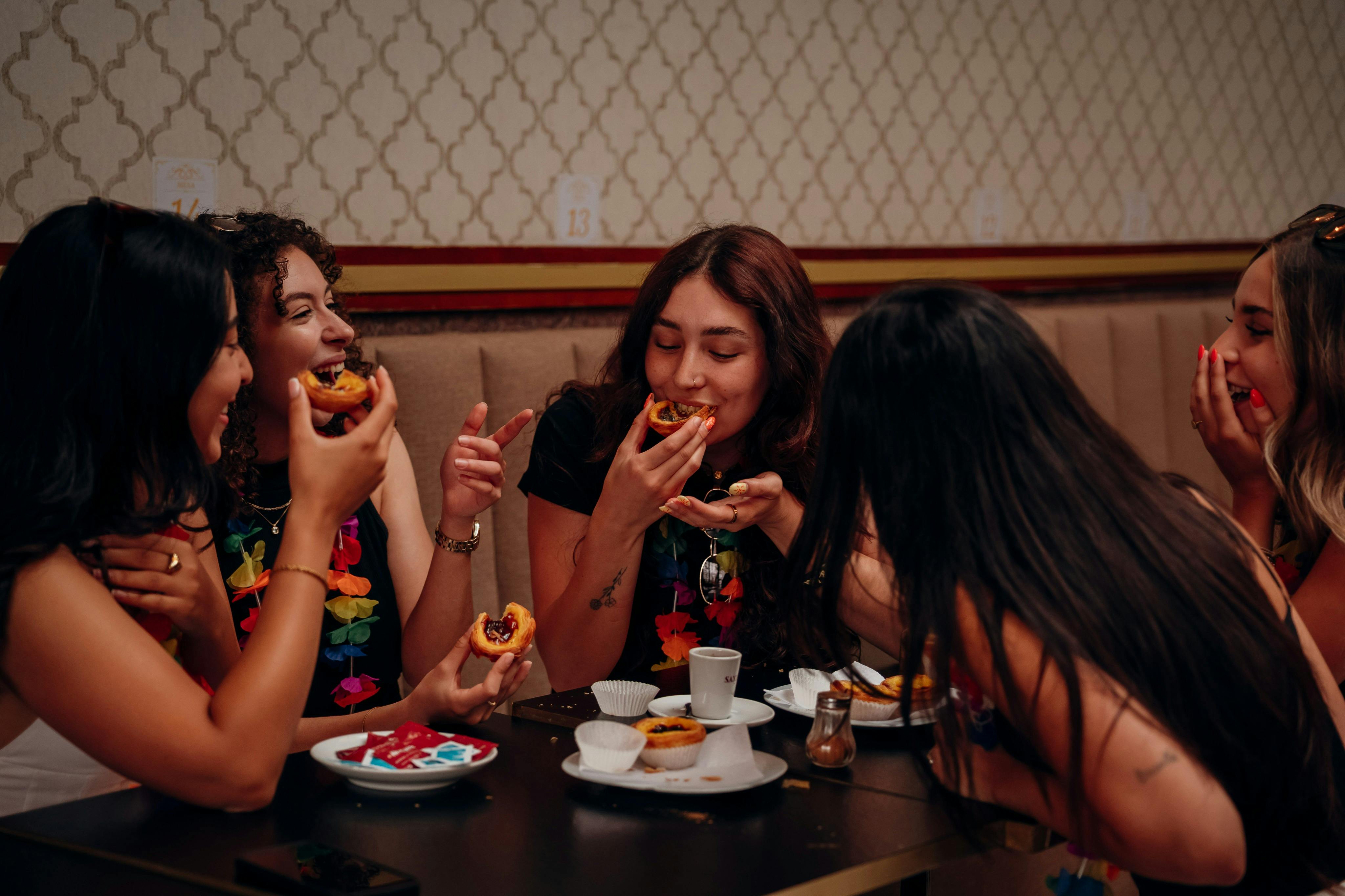 Drei mit bunten Blumenkränzen geschmückte Frauen essen fröhlich Gebäck und unterhalten sich an einem Tisch mit Tellern und Tassen in einem gemütlichen Café.