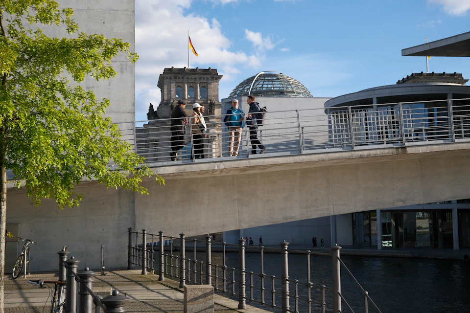 A group of people standing on a bridge with the Reichstag building and its glass dome visible in the background.