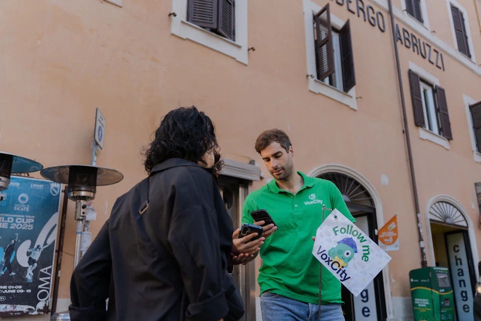 A person in a green "VoxCity" shirt holding a sign speaks with another individual near a building with arched windows.