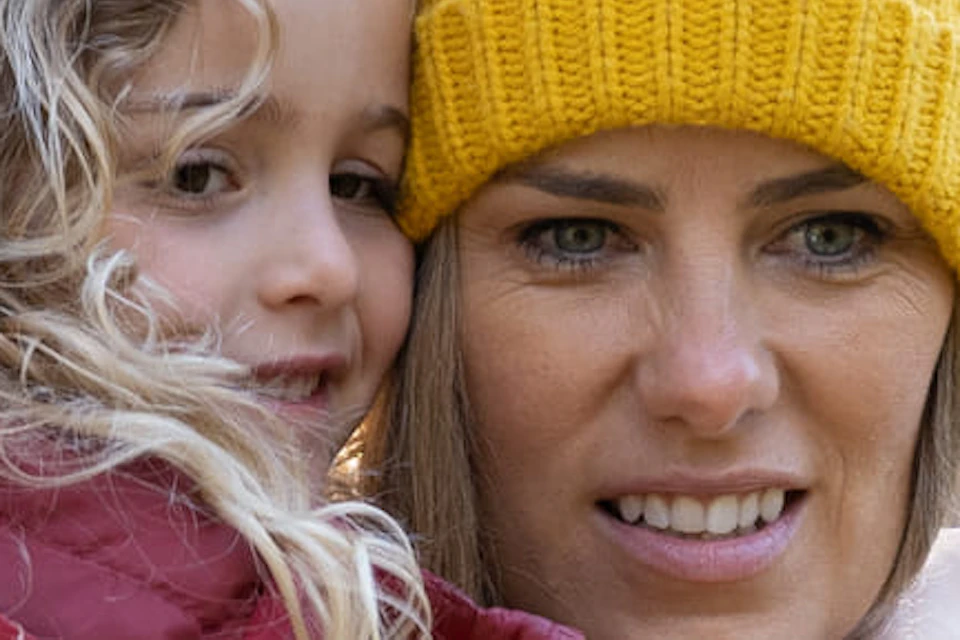 A woman in a yellow beanie and a child in a red jacket stand near a koala in a zoo setting with trees in the background.