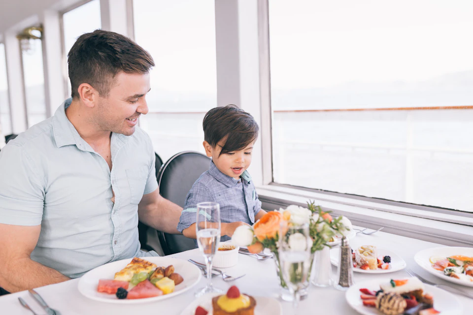 A man and a young boy sit at a table with plates of food and flowers by a window, smiling and engaging with each other.