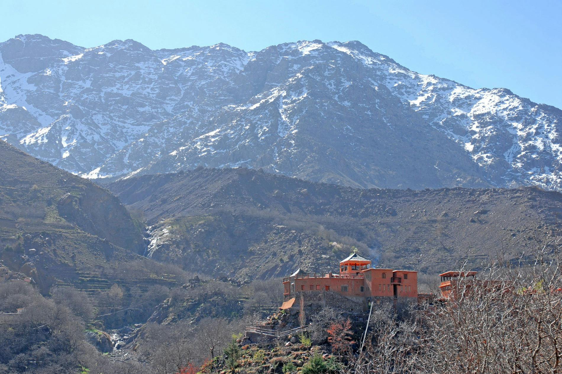 Красное здание расположено на склоне холма с заснеженными горами на заднем плане под ясным голубым небом.