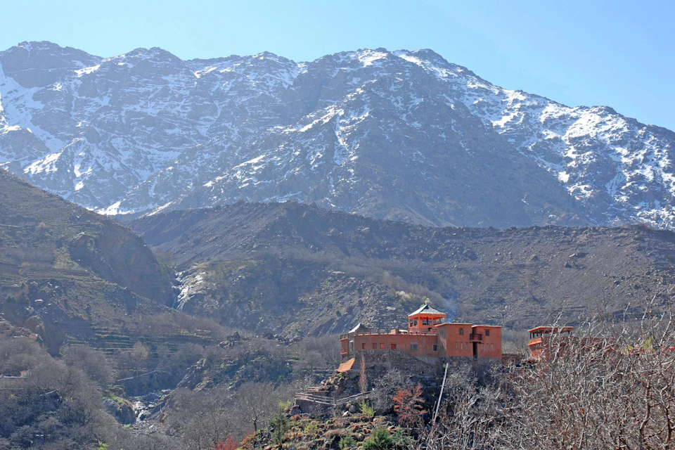 A red building is situated on a hillside with snow-capped mountains in the background under a clear blue sky.