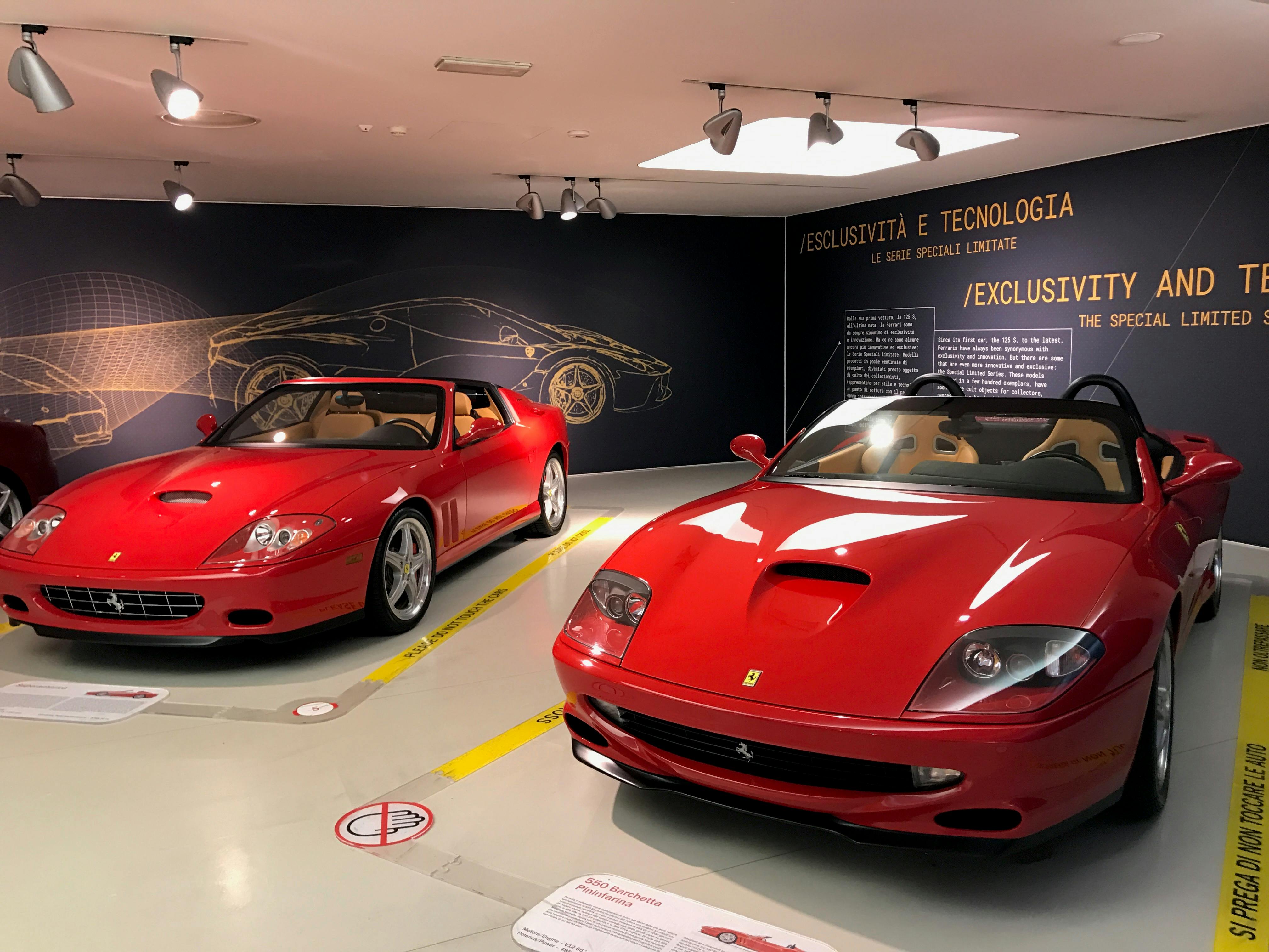 Δύο κόκκινα κάμπριο Ferrari που εμφανίζονται σε έναν εκθεσιακό χώρο με ενημερωτικούς πίνακες στον τοίχο πίσω τους.