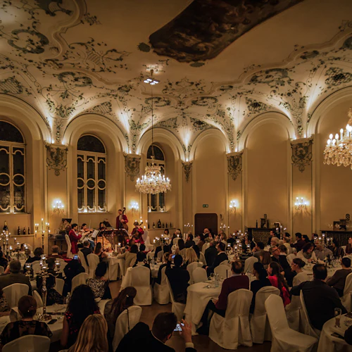 Gäster i elegant klädsel sitter vid runda bord i en stor sal med högt i tak, ljuskronor och utsmyckad inredning och lyssnar på musiker.