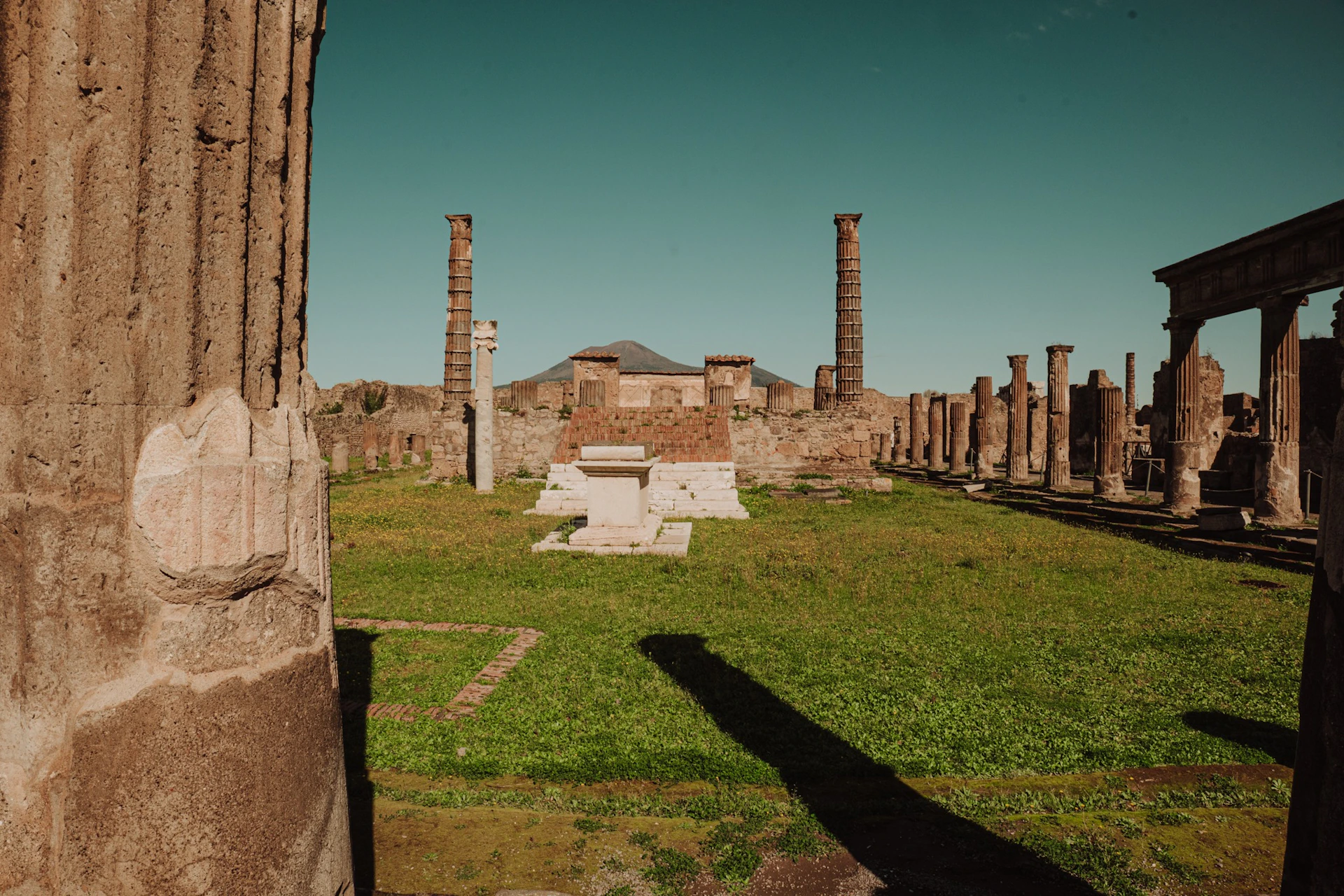 Ruins of an ancient structure with stone columns and a central altar, set against a clear sky and distant mountain.