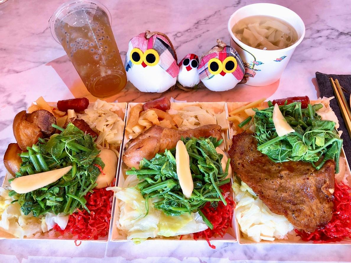 A meal consisting of three plates of food with greens, meat, and sides, accompanied by a drink, soup, and decorative figurines.