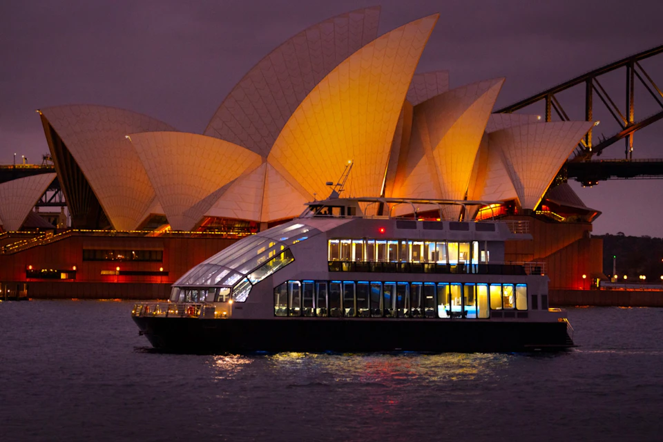A modern ferry with illuminated windows passing in front of the Sydney Opera House at dusk.