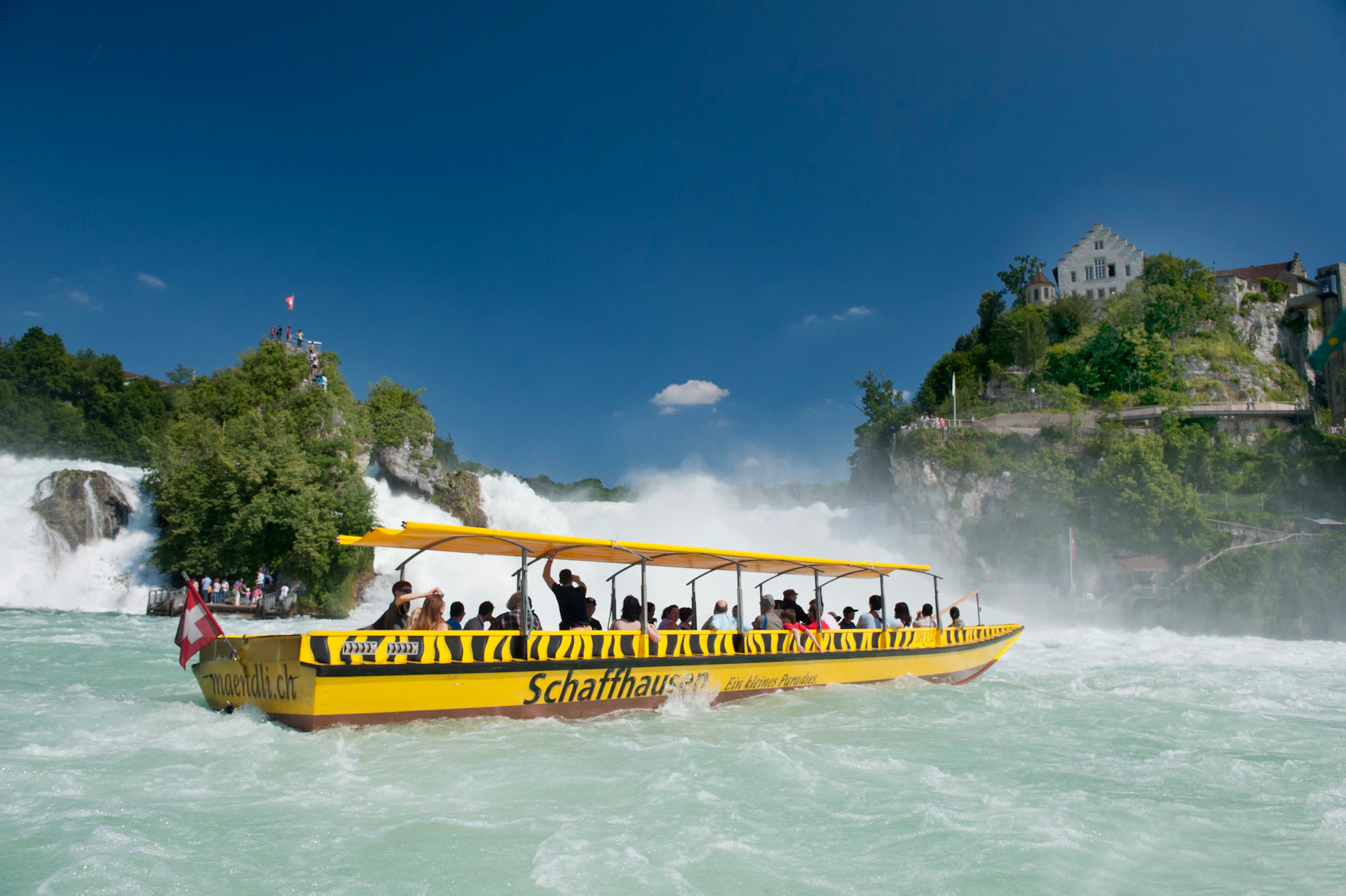 People on a yellow boat with "Schaffhausen" written on the side, approaching a mist-covered waterfall under a clear blue sky.