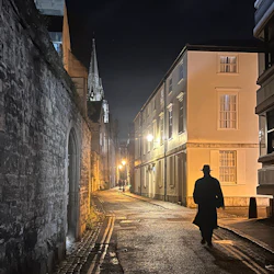 A dimly lit cobblestone street at night, with a silhouette of a person in a hat and coat walking away under streetlights.