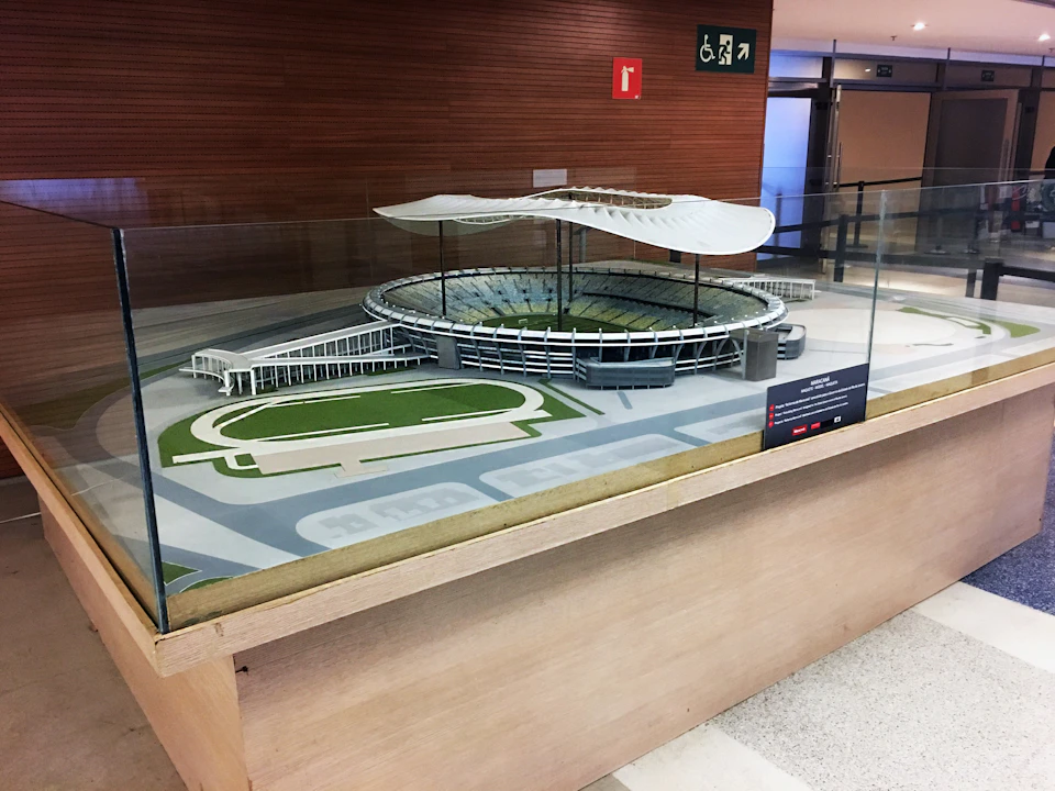 A detailed scale model of a stadium with seating, a track, and a unique wavy roof, enclosed in a glass display case indoors.