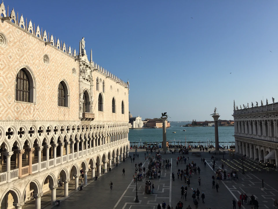 Gothic palace next to a canal with columns and statues, crowded square below, and distant buildings across the water under a clear sky.