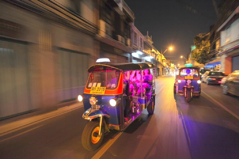 Two tuk-tuks with illuminated interiors drive down a city street at night, with blurred buildings in the background.