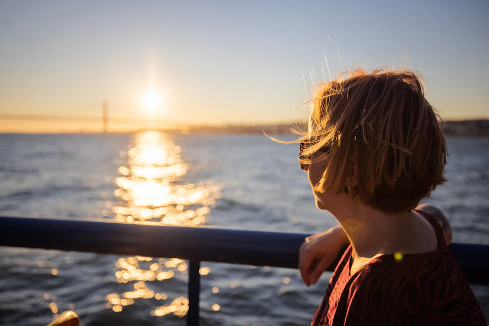 A person with short hair stands at a blue railing, looking at a sunset over a body of water.
