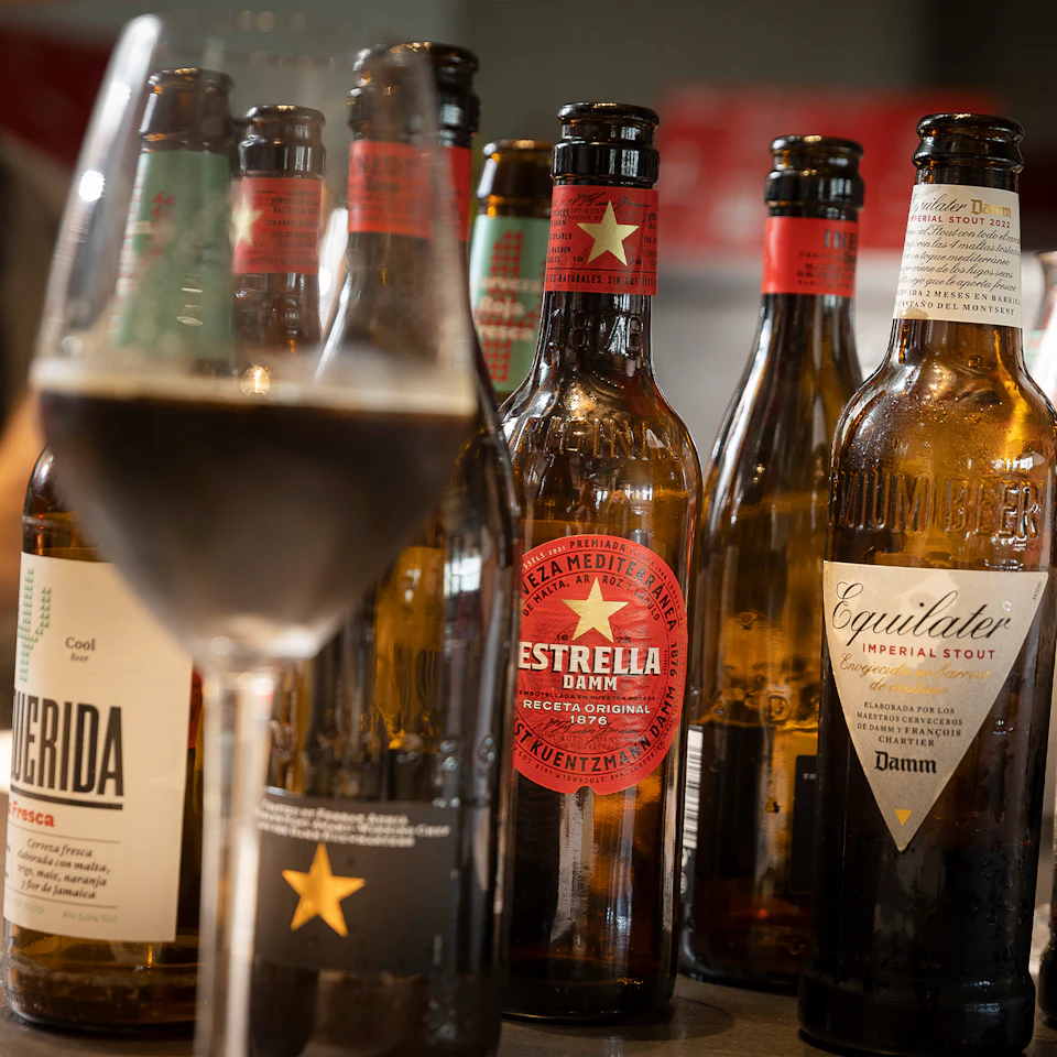 Several beer bottles with various labels, including Estrella Damm and Equilater, next to a full glass in the foreground.