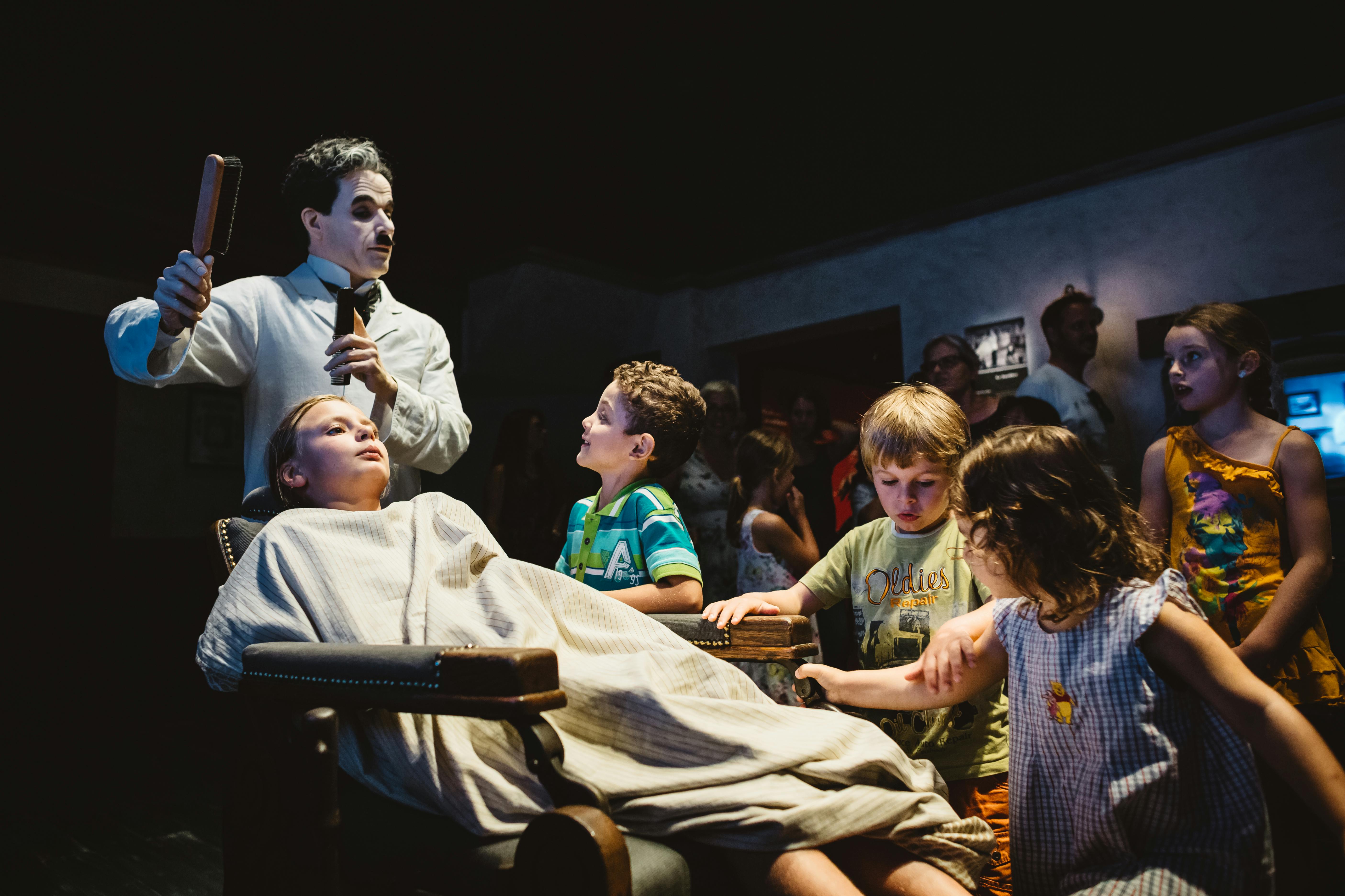 Děti se shromáždí kolem osoby pokryté prostěradlem, s postavou připomínající klauna nebo performera v bílém make-upu a černém oděvu.