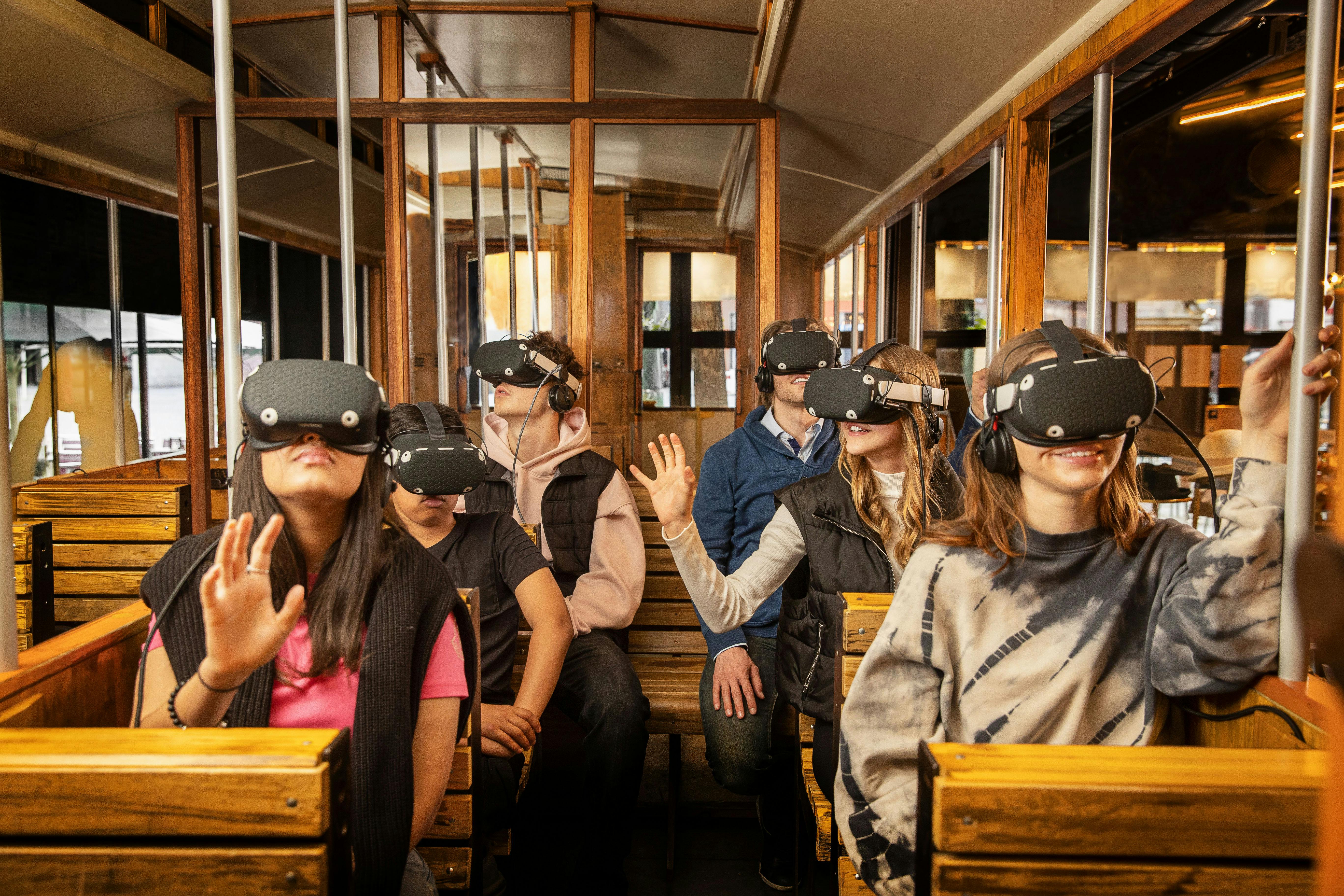 Cestující v dřevěné tramvaji s náhlavními soupravami pro virtuální realitu, někteří se natahují a usmívají.
