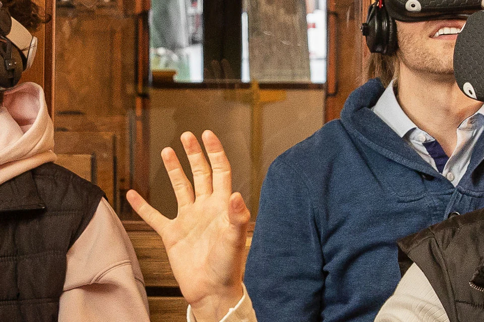 Passengers on a wooden tram wearing virtual reality headsets, with some reaching out and smiling.