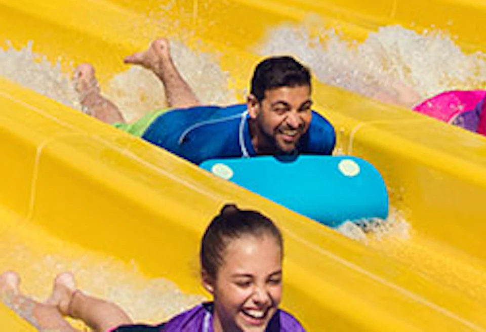 People on blue mats racing down adjacent yellow water slides at a colorful water park.