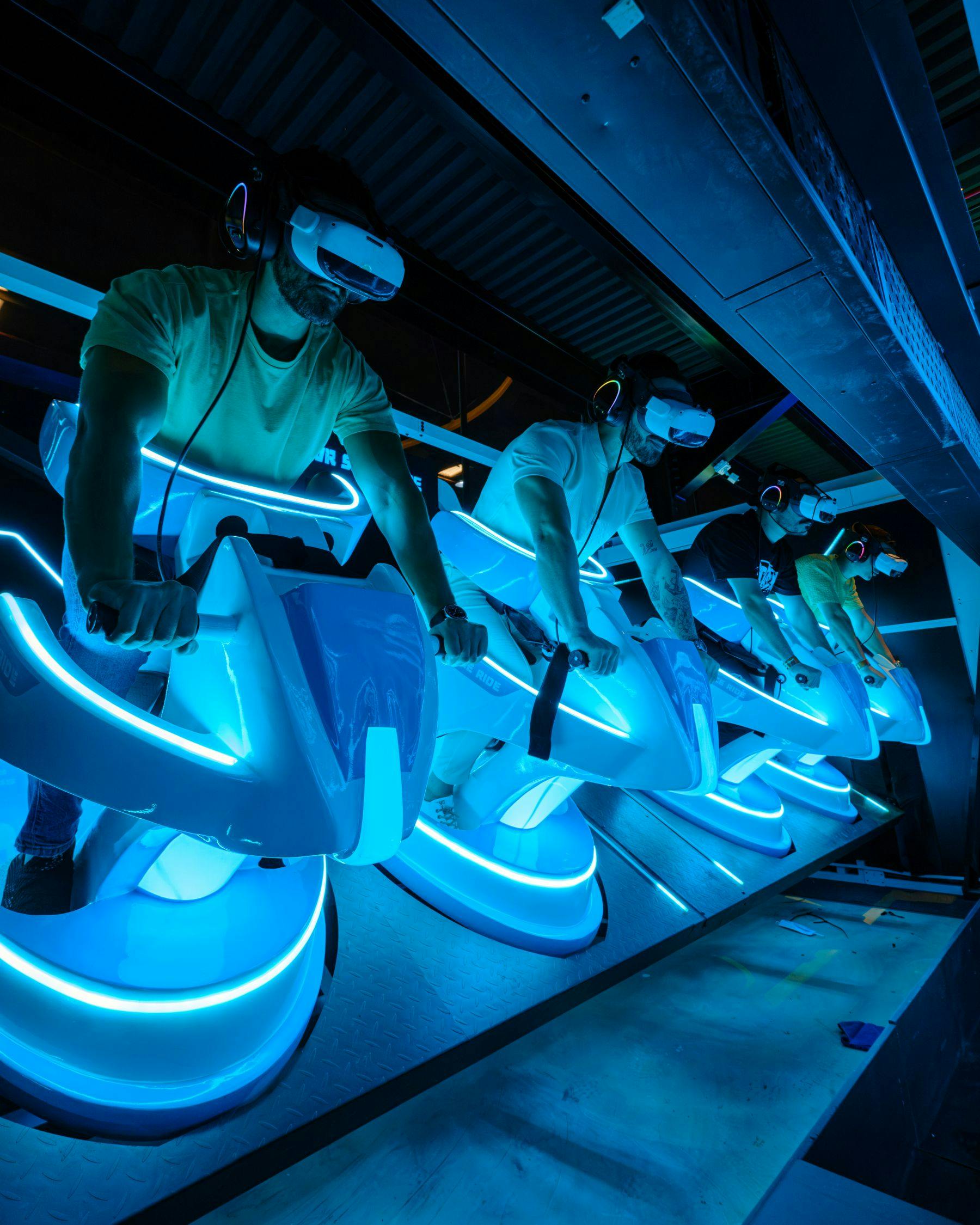 Άτομα που χρησιμοποιούν ακουστικά εικονικής πραγματικότητας ενώ κάθονται σε φωτιζόμενους φουτουριστικούς προσομοιωτές ποδηλάτων σε περιβάλλον με φως νέον.