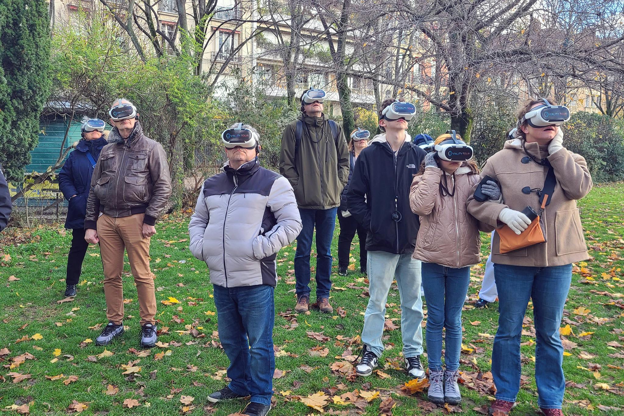 Un gruppo di persone all'aperto che indossano cuffie VR, in piedi su un prato circondato da alberi e foglie cadute.