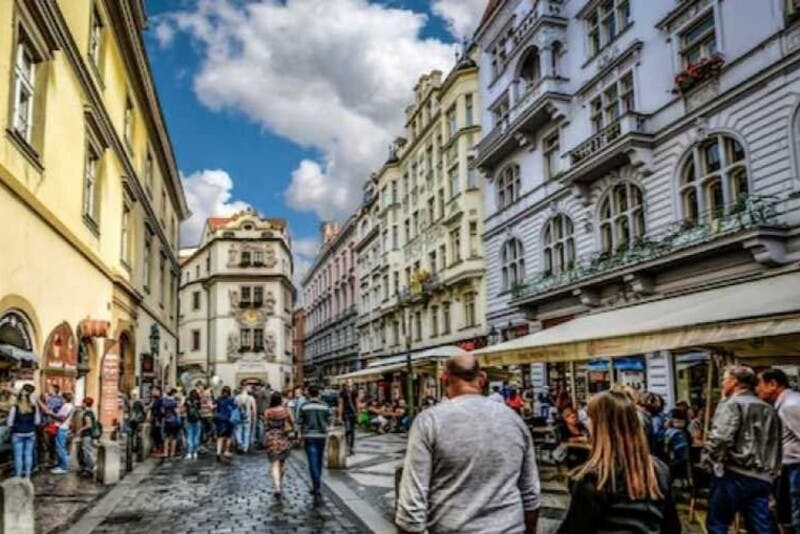 部分的に曇った空の下、豪華な建物や屋外カフェが並ぶヨーロッパの通りを歩く歩行者。