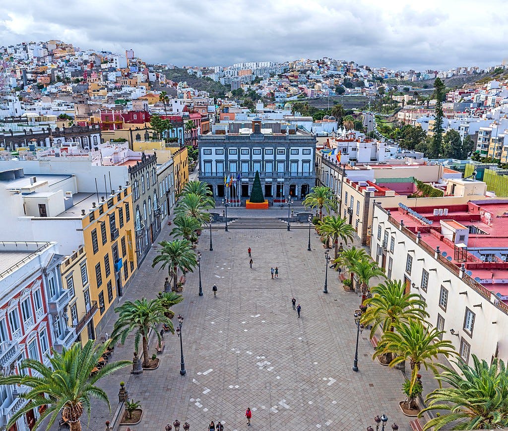 Vista aérea de uma grande praça com palmeiras, edifícios coloridos e pessoas a passear; uma árvore de Natal junto a um edifício cinzento.