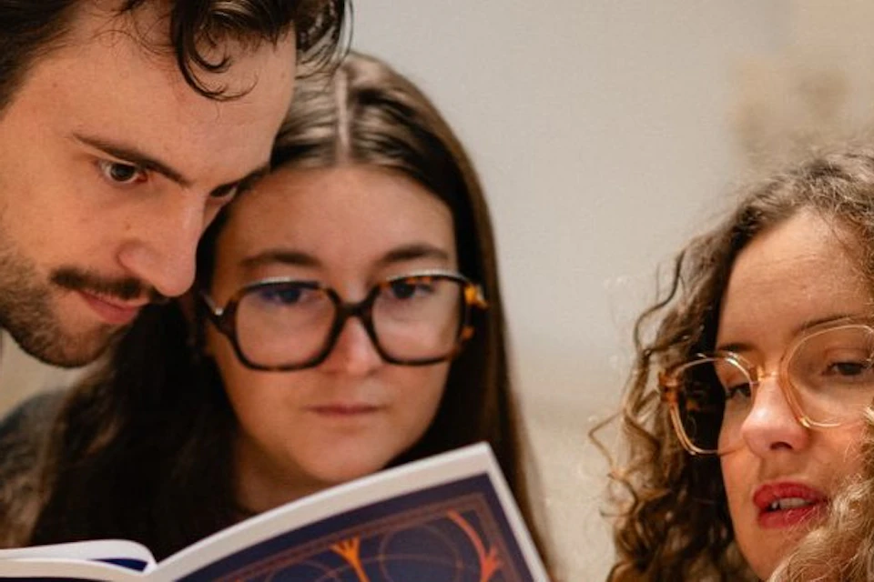 Three people are looking at an open booklet entitled 'Mazarin'; they appear to be discussing it or examining it closely.