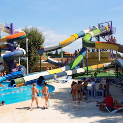Människor njuter av en vattenpark med färgglada rutschkanor, en pool och loungeområden under en solig himmel.