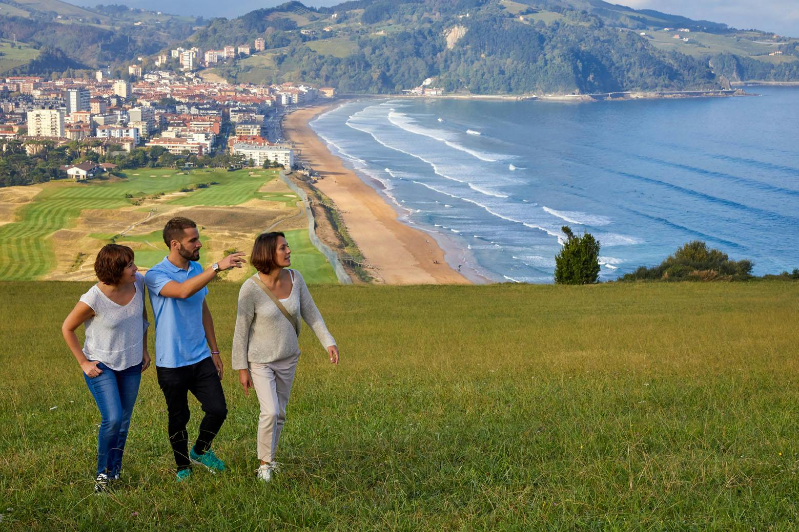 Three people walking on a grassy hill overlooking a coastal town with a beach, buildings, and mountainous background.