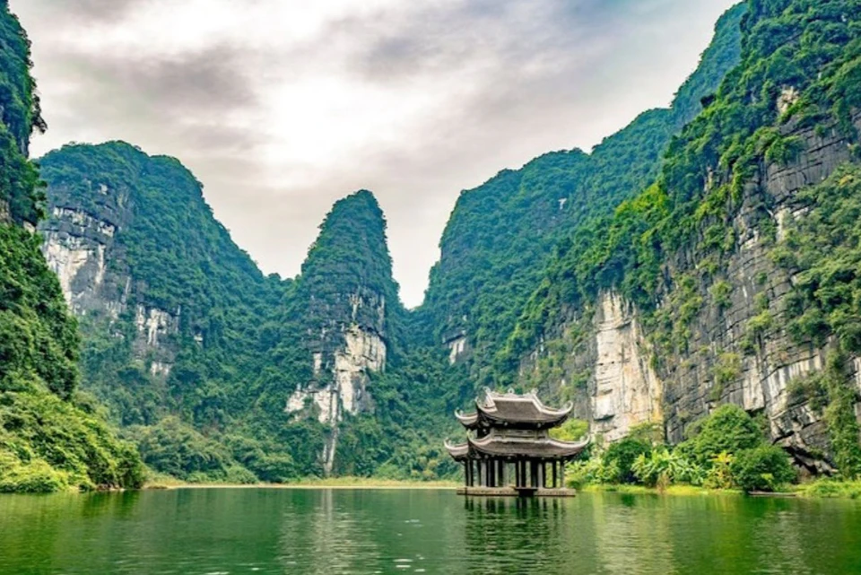 Traditional pavilion on water surrounded by tall, lush green mountains under a cloudy sky.