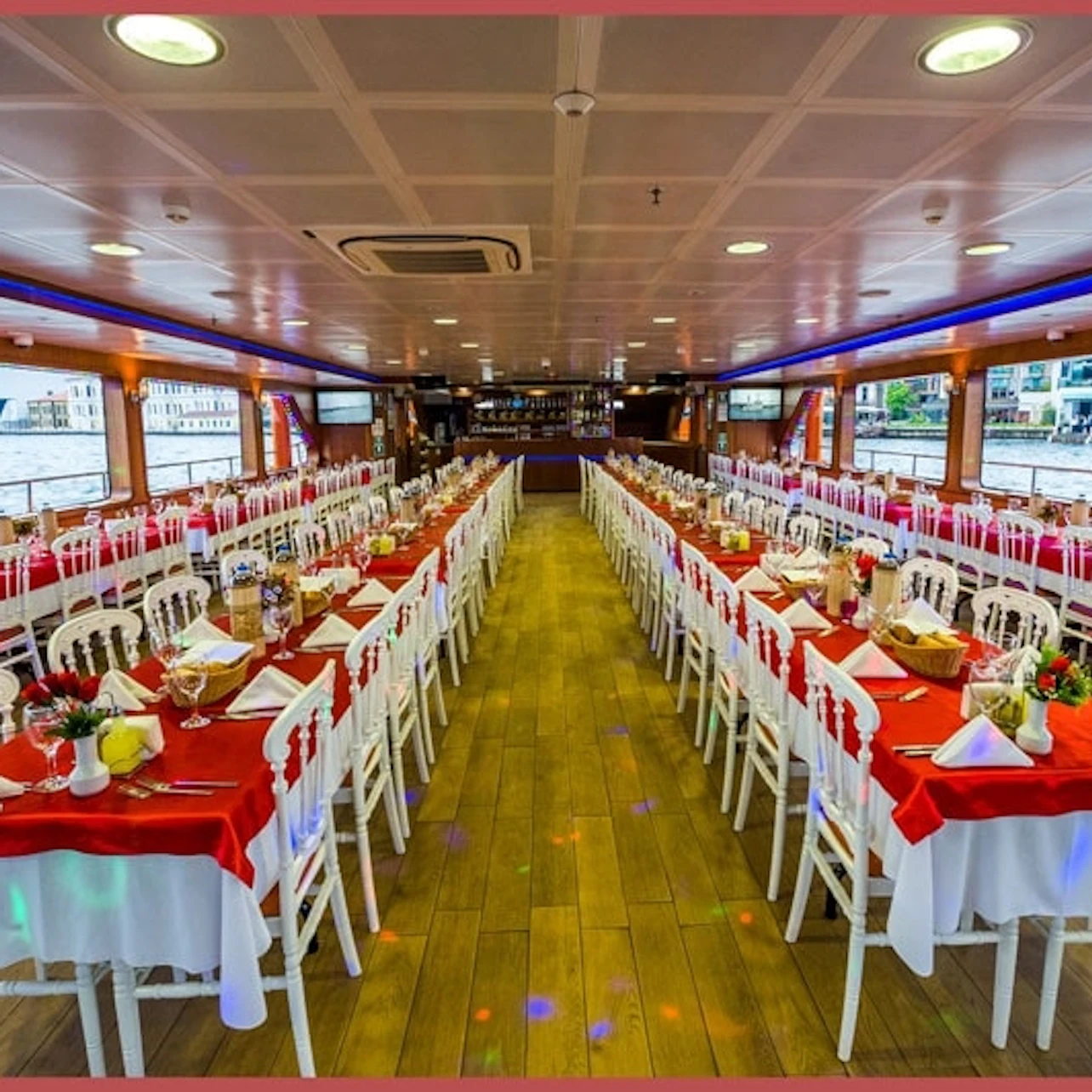 A large, elegantly set dining area on a boat with red and white table settings, arranged in long rows with windows on both sides.