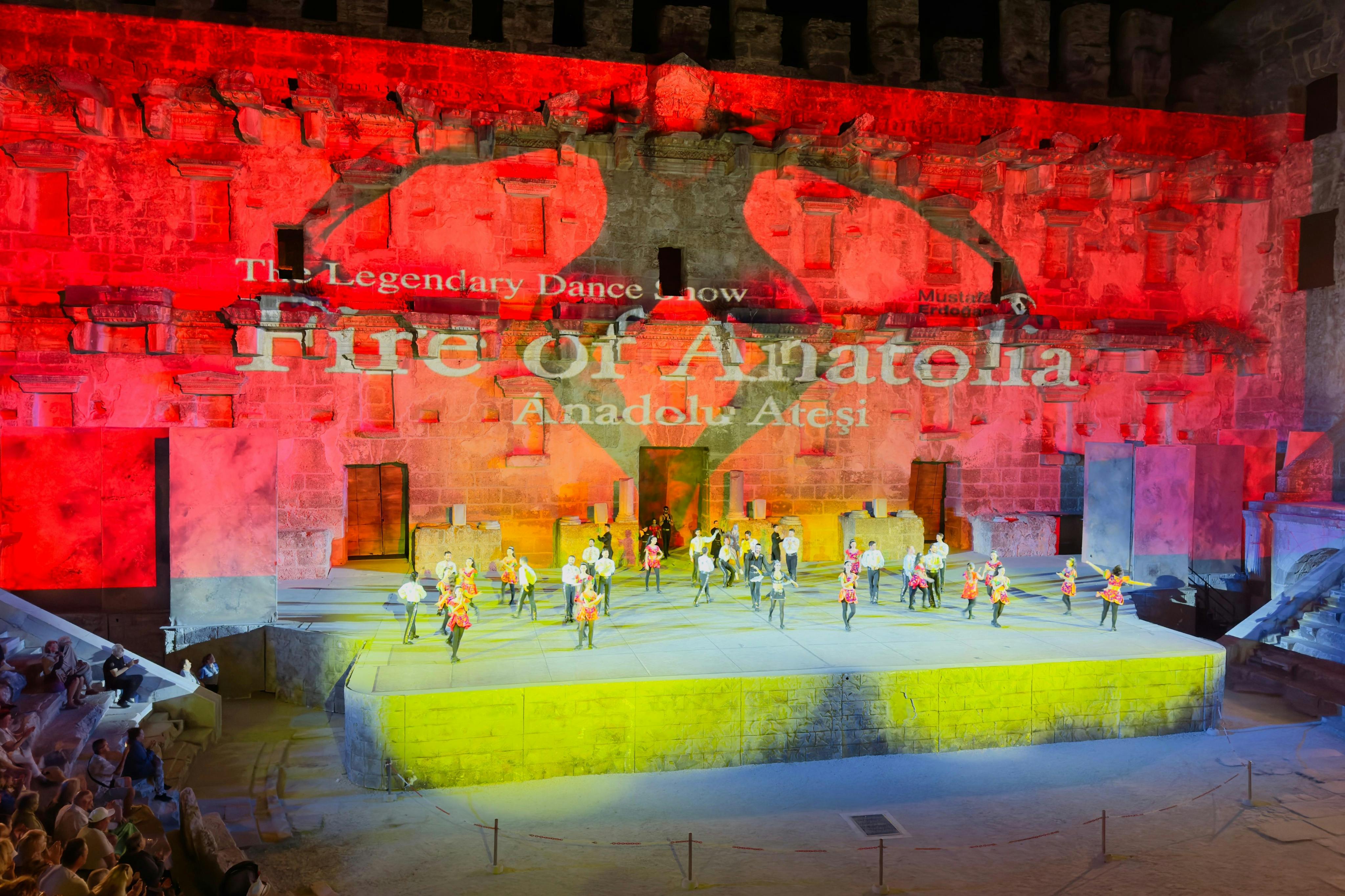 Танцевальный спектакль на освещенной сцене с фоном «Огонь Анатолии». Исполнители носят яркие костюмы.