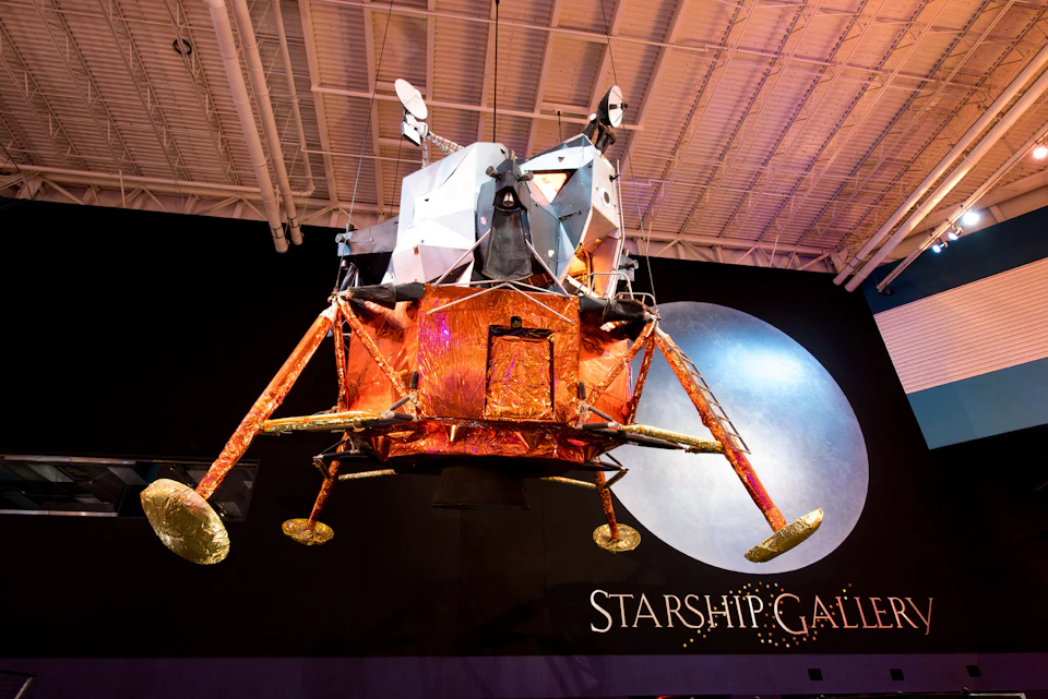 A model of a lunar lander is suspended from the ceiling with "STARSHIP GALLERY" written on the wall behind it.