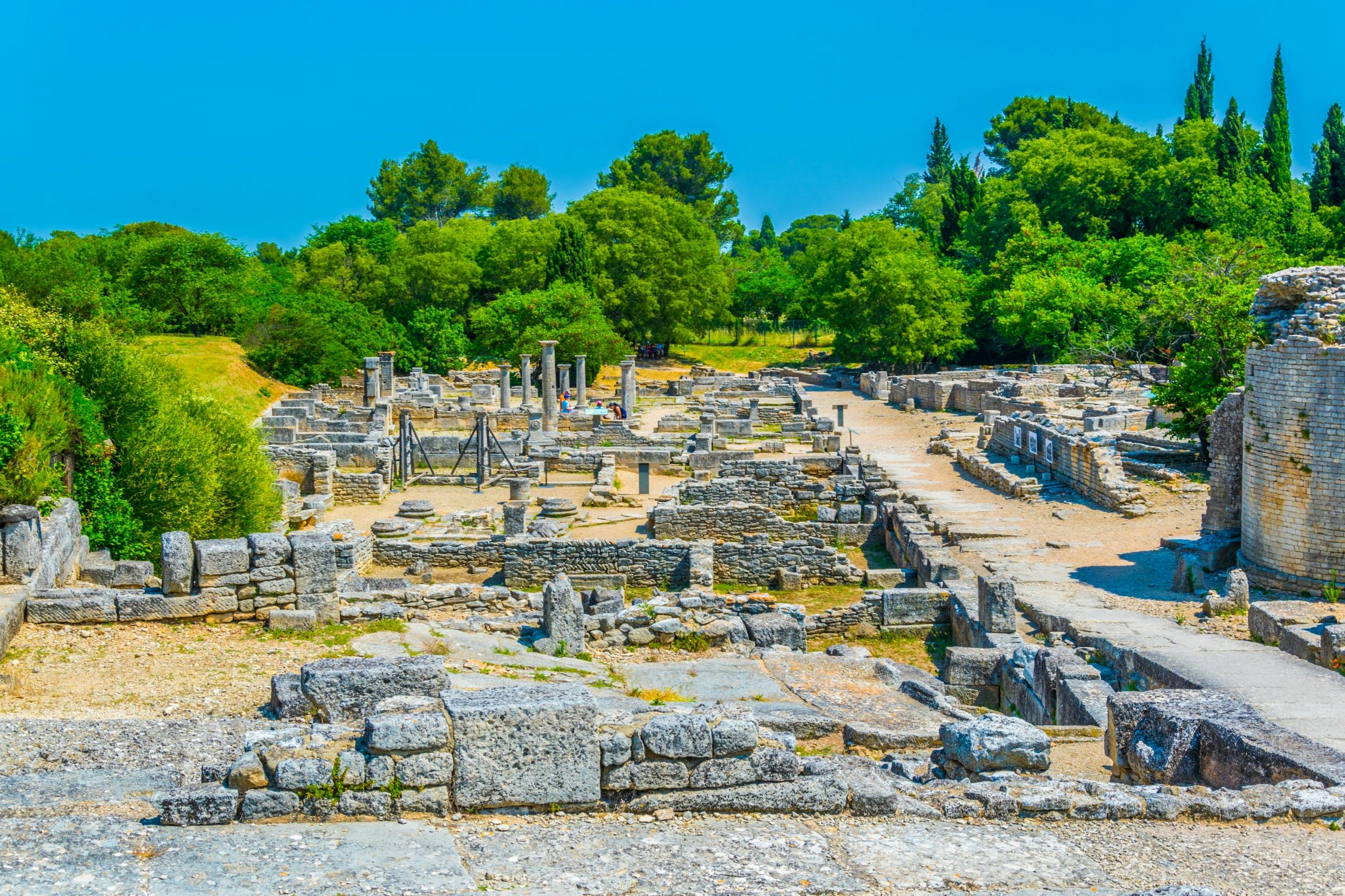 Ancient stone ruins with columns and walls, surrounded by green trees under a clear blue sky.