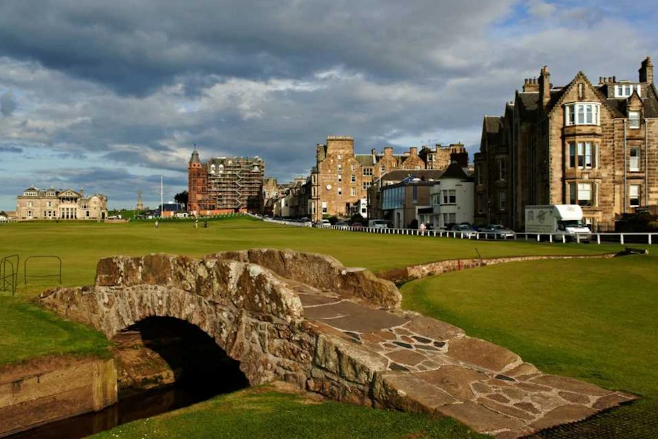 A stone bridge over a grassy area, with a historic town and cloudy sky in the background.