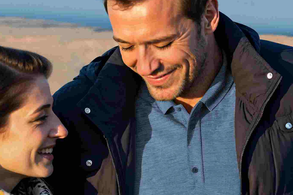 Three people in jackets talking and smiling near a railing overlooking a scenic, rocky landscape under a partly cloudy sky.