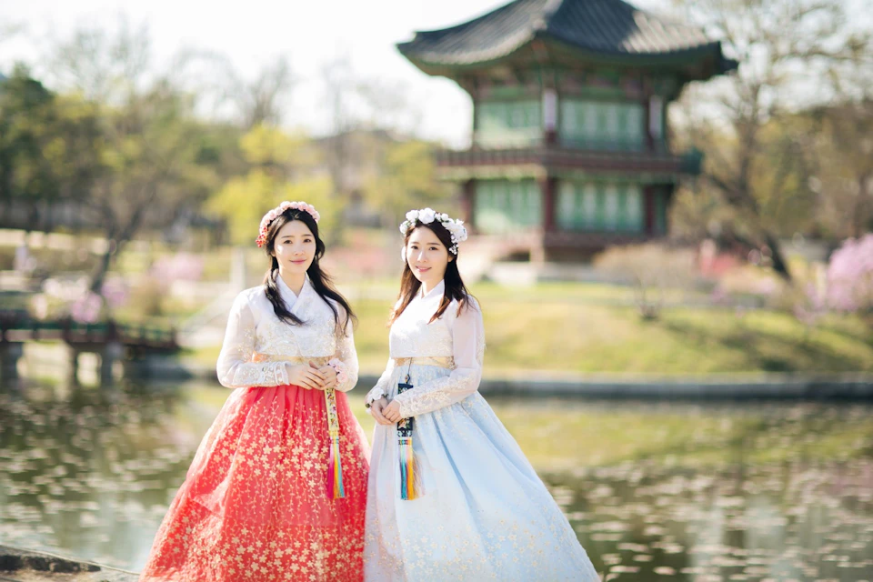 Two women in traditional Korean hanbok dresses, one in red and one in blue, standing outdoors near a pond with a pavilion.