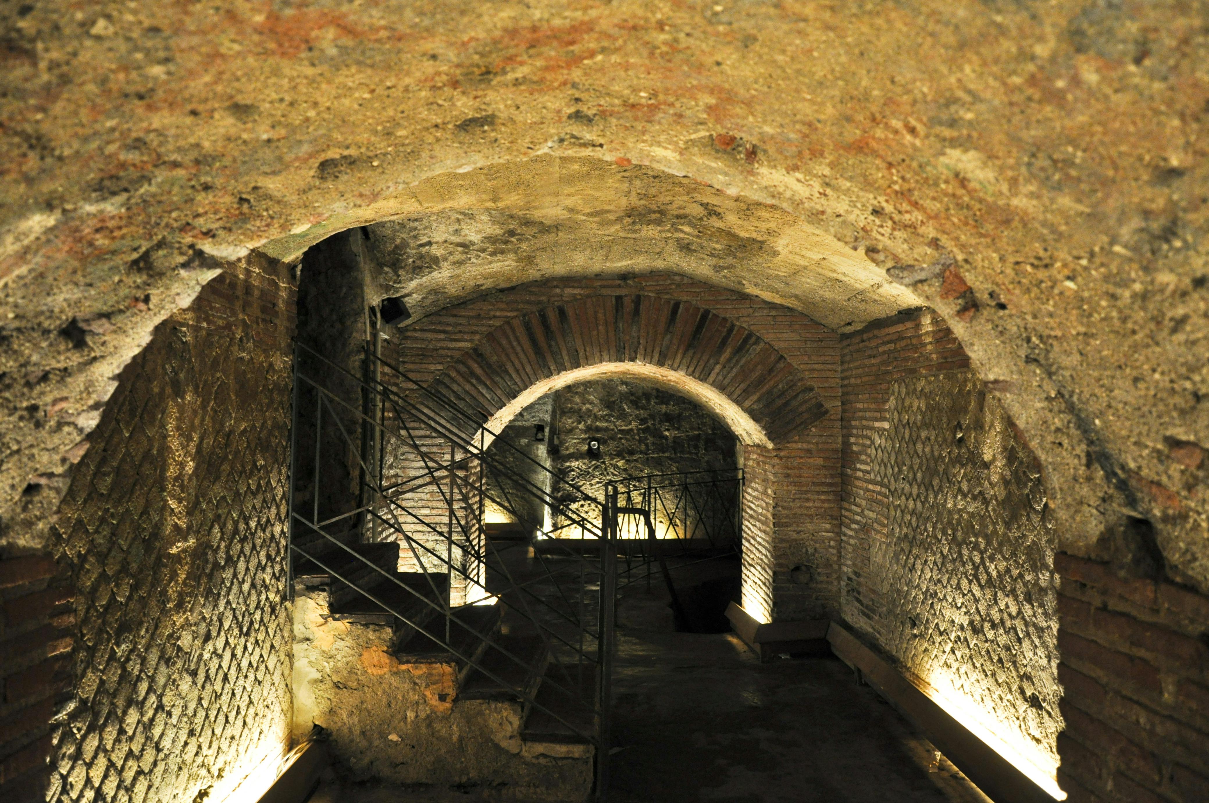 Un passage souterrain faiblement éclairé avec des murs en pierre, des arches voûtées et un escalier métallique menant vers le haut.