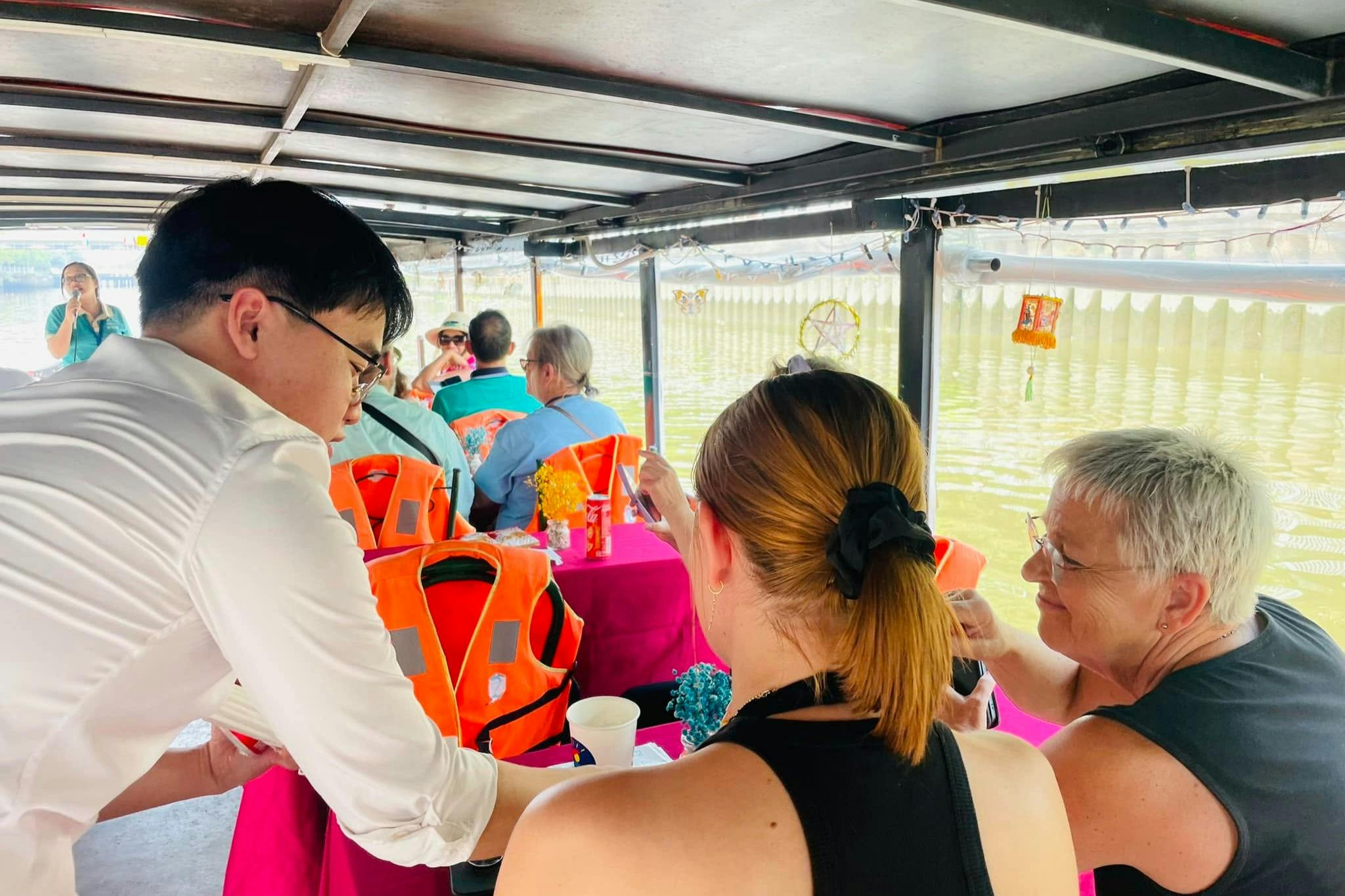 Menschen sitzen an Tischen auf einem Flussschiff, einige tragen orangefarbene Schwimmwesten. Ein Kellner im weißen Hemd kümmert sich um die Passagiere.