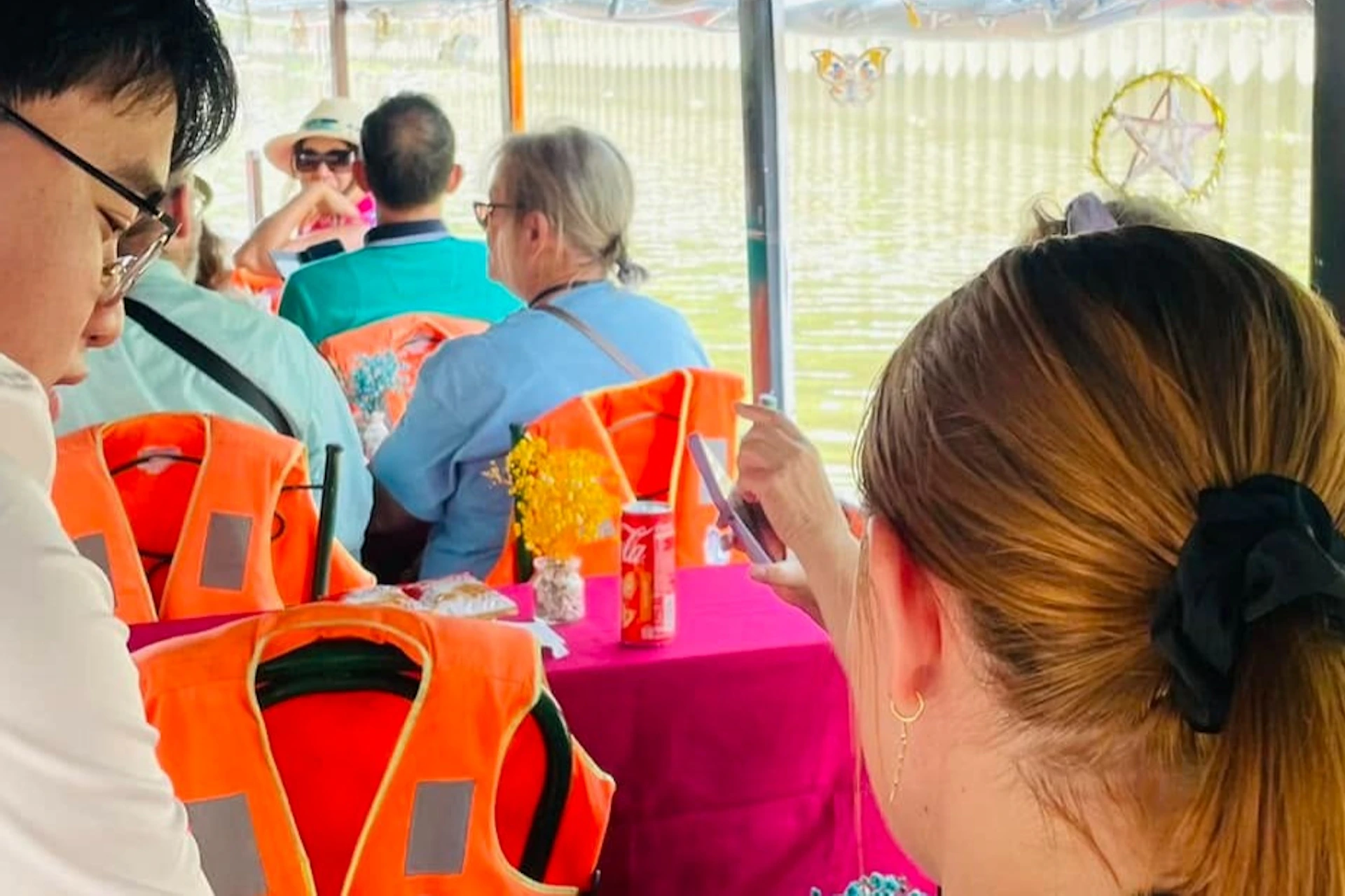 People seated at tables on a riverboat, some wearing orange life jackets. A server in a white shirt attends to passengers.